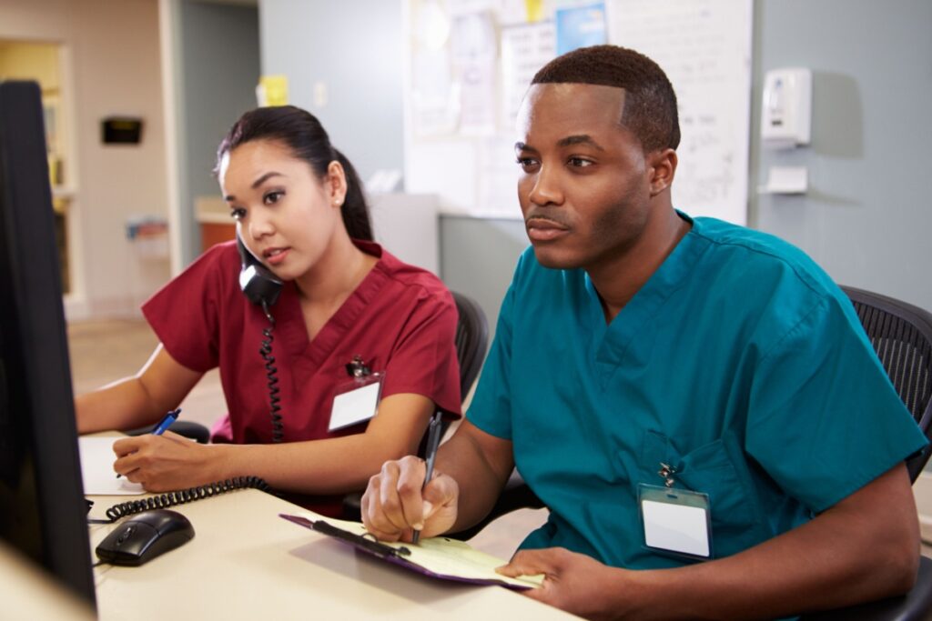 Two healthcare workers in scrubs sit at a desk, reviewing notes and speaking on the phone in a clinical setting. 