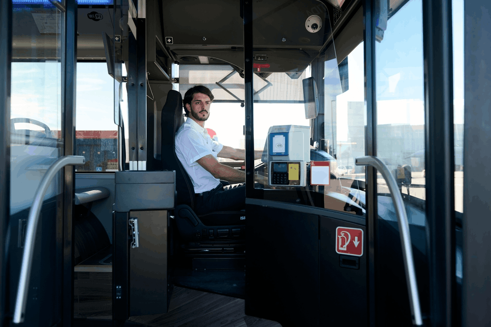 City bus driver seated in a bus cab behind a glass partition, hands on the wheel.