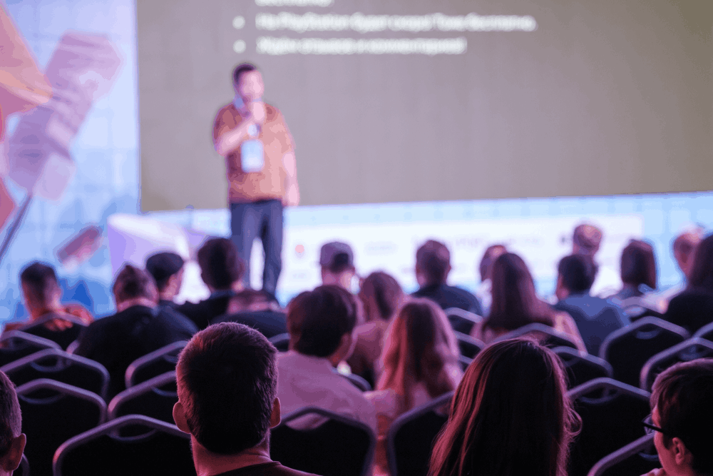 Audience seated in a conference room watching a blurred speaker present onstage in front of a large screen. 