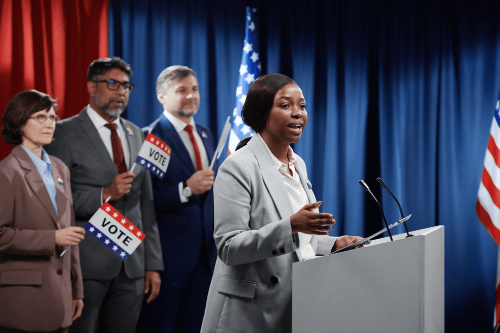 A political candidate speaking at a podium with supporters holding vote signs, illustrating what political parties do during elections. 