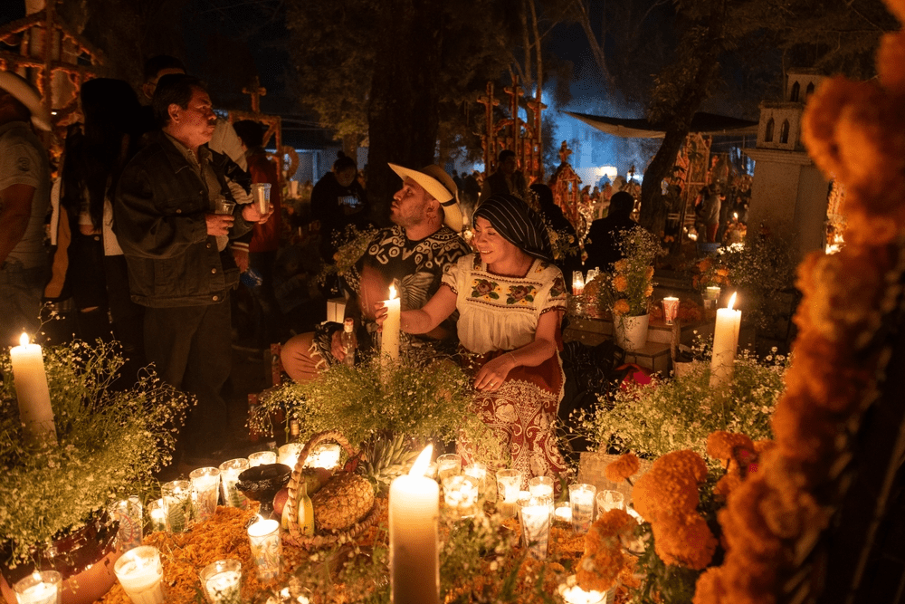 People gather in a candlelit cemetery adorned with marigolds and offerings during a Día de los Muertos celebration. 