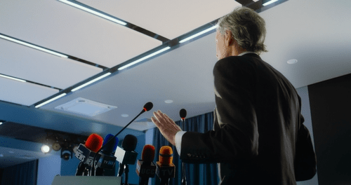 A politician gives a speech on the campaign trail, surrounded by microphones. 