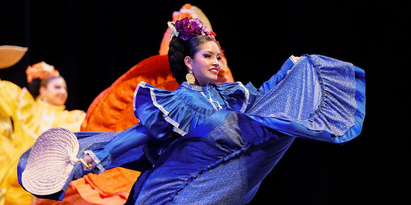 A woman in a blue folklorico dress dances while holding a fan on stage, with colorful dancers in the background. 