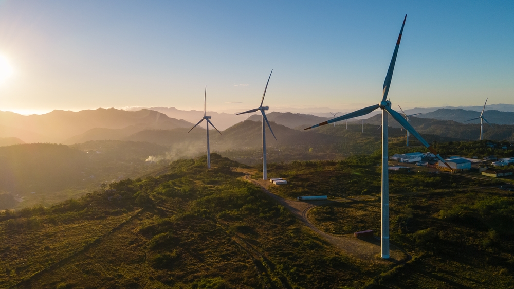 Aerial view of wind turbines on a green hillside at sunset, with mountains in the background.