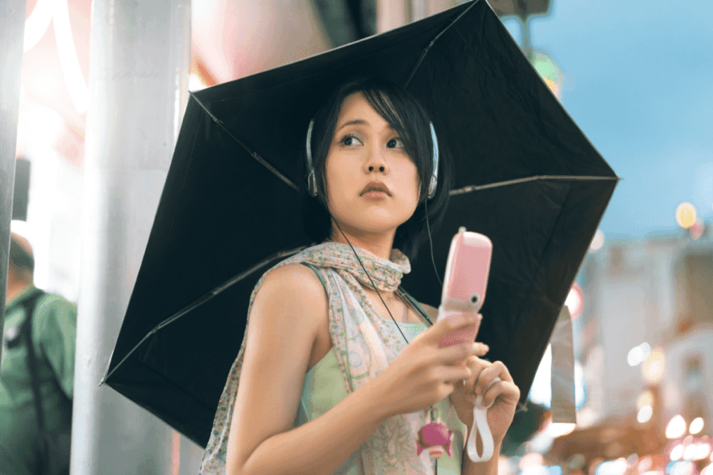 Woman standing on a city street corner, holding a pink flip phone in one hand and a black umbrella in the other.