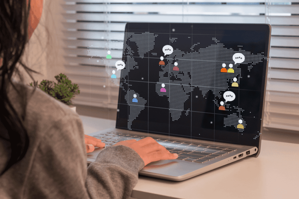 A student types on a laptop showing a world map with digital icons representing people and speech bubbles, symbolizing global communication and intercultural connection. 