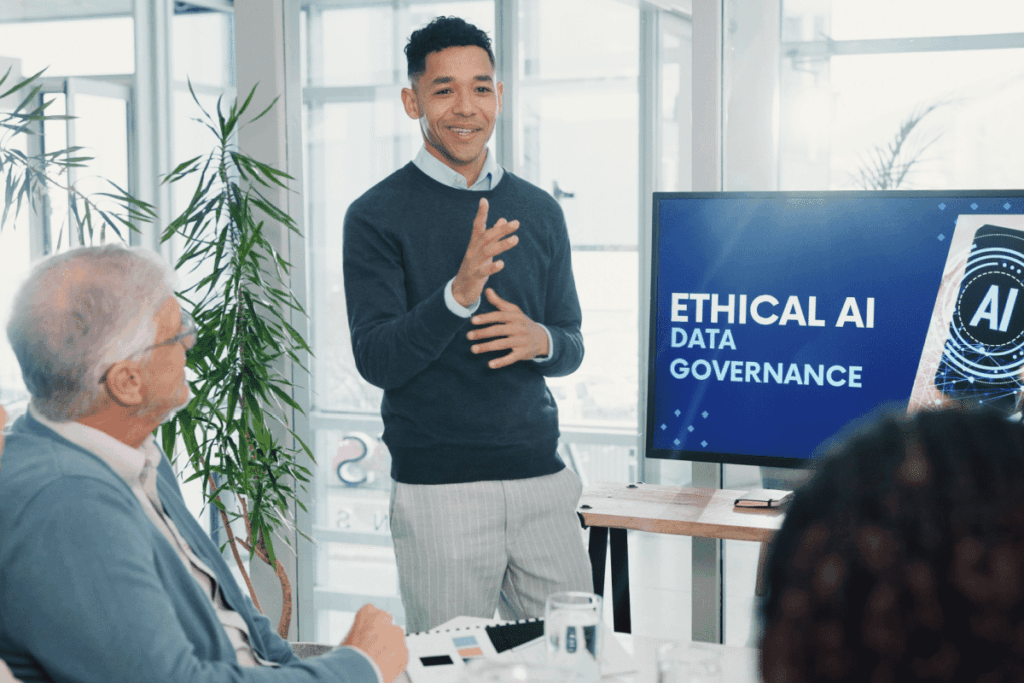 A man stands and smiles while giving a presentation on ethical AI to a group of seated people in a meeting room. The presentation on his screen reads “Ethical AI Data Governance.” 