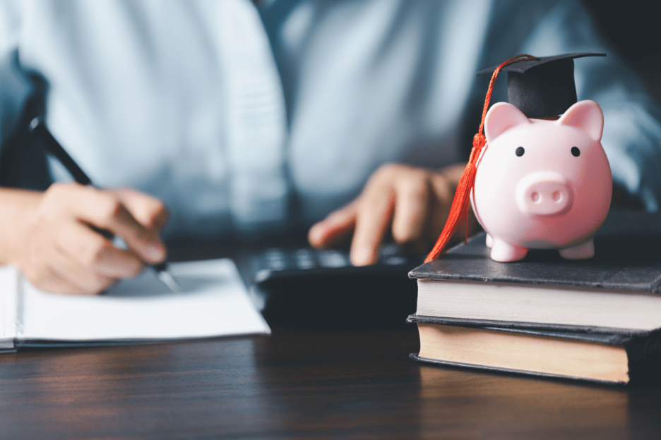 A pink piggy bank wearing a graduation cap sits on stacked books while a person calculates tuition costs in the background.
