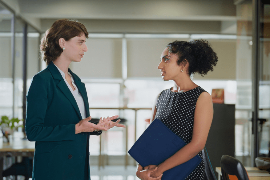 Two professional women have a friendly conversation in a modern office. One wears a dark green blazer and gestures while speaking, while the other holds a blue folder and listens attentively.