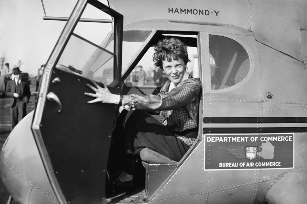 Amelia Earhart sits in the pilot’s seat of a single-engine plane, holding open the door. 