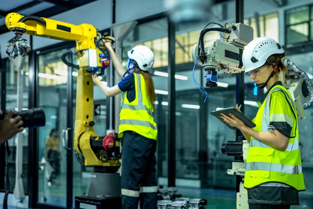 Two engineers in safety gear work with robotic arms in a manufacturing facility. One adjusts equipment while the other reviews data on a tablet.