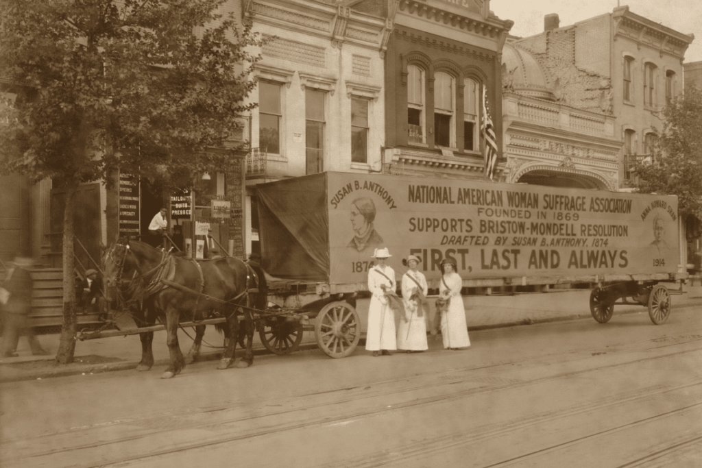 Sepia-toned historical photo of women wearing white dresses, standing beside a suffrage banner during the early American women’s suffrage movement.