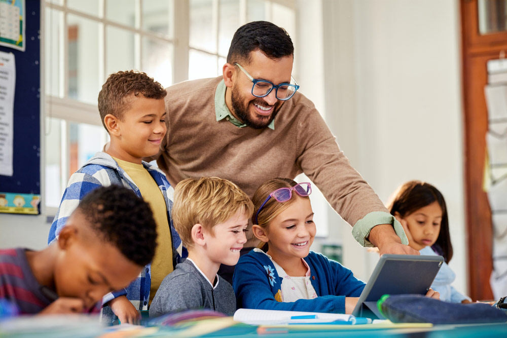 Male teacher helping diverse elementary students working with tablets and notebooks at a classroom table. 