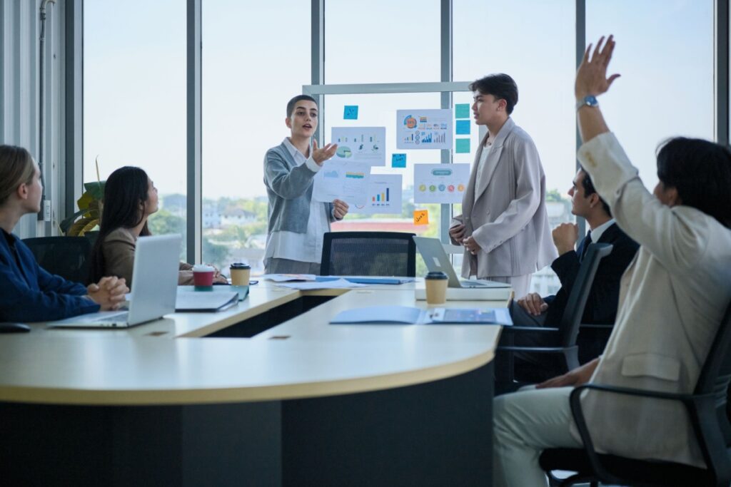 Two professionals present charts on a glass wall to seated colleagues in a conference room, one raising a hand.