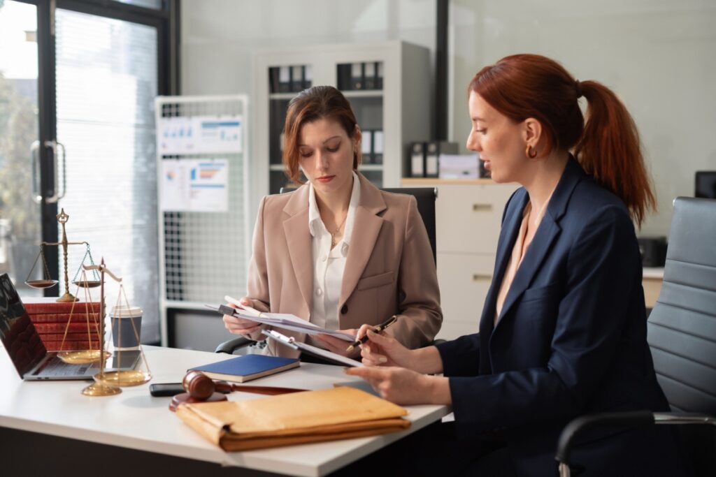 Two women in business attire review documents at an office desk. 