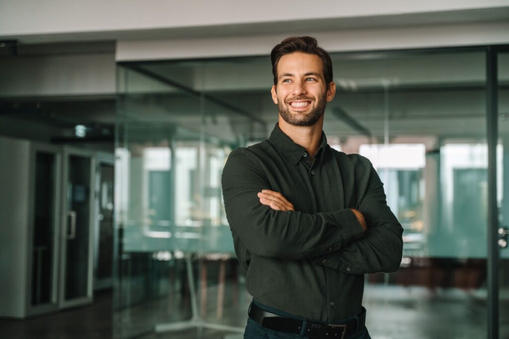 A smiling man stands with arms crossed in modern glass-walled office. 
