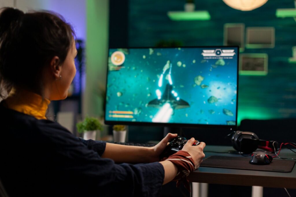 Gamer using a controller at a desk, playing a space-themed video game on a computer screen in a dimly lit room.