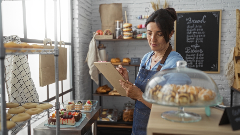 Bakery owner reviewing notes in her shop among baked goods and customer orders. 