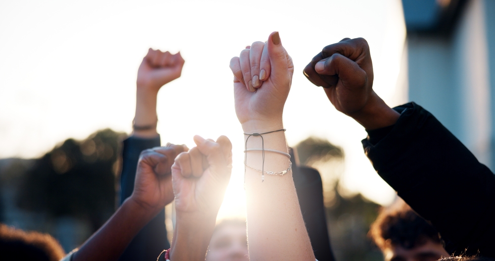 Several raised fists and clasped hands lifted in solidarity against a bright sky.  